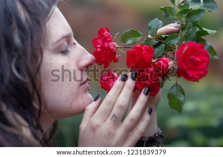 Beautiful young individual, eccentric woman, with attractive dreadlocks, piercing and tattoo, who smells with eyes closed at red roses in the garden park
