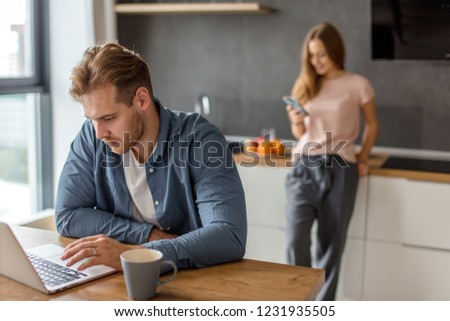 nice guy typing a message in the kitchen room ,attractive woman using gadget in background.
