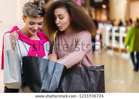 Two satisfied attractive female shoppers talking after shopping trip, discussing their purchases, standing in the hall of shopping mall.