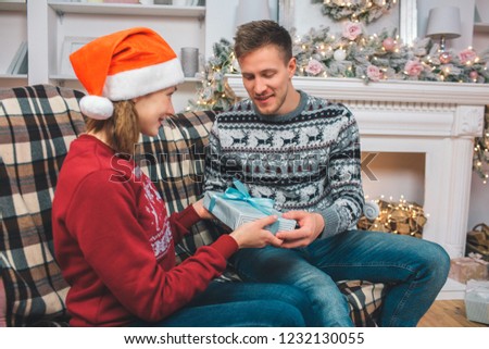 Picture of young man sitting on sofa. He gives packed blue box to young woman. She is excited and happy. Man is happy. They holds gift together.