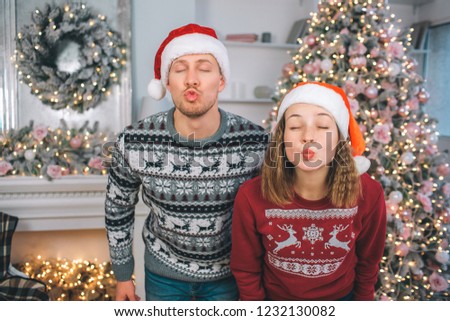 Funny picture of young man and woman stands and poses. They keeps eyes closed. People's lips are in kiss shape. There are Christmas tree and fireplace behind them.