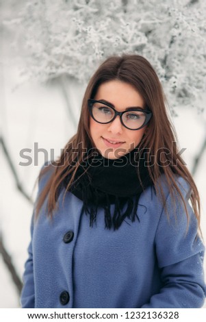 Portrait of happy young woman wearing winter hat and scarf stands next to the Christmas tree. black glasses