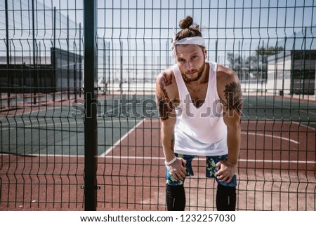 Young man with white headband  on his head and tattoos on his arms dressed in the white t-shirt, black leggings and blue shorts stands next to the playground fence outside on a sunny day