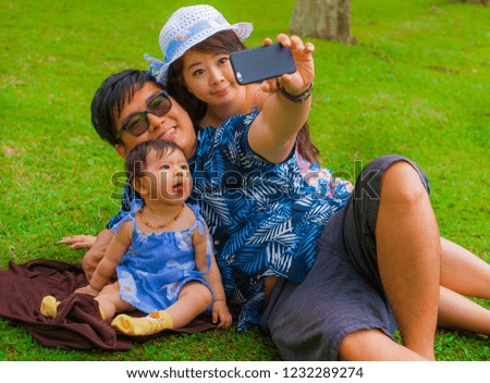young happy and loving Asian Japanese family with parents and sweet baby daughter at city park together with father taking selfie pic with mobile phone for uploading on internet social media