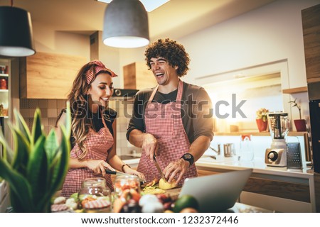 Young happy couple is enjoying and preparing healthy meal in their kitchen and reading recipes on the laptop. Royalty-Free Stock Photo #1232372446