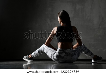 Young sporty woman after gymnastics stretching fitness exercises workout sitting on floor backside view on dark wall background 