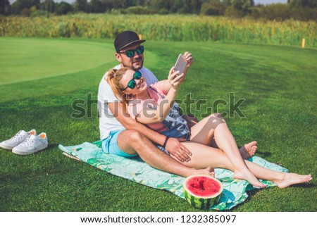 Young man and his pregnant wife eating watermelon in park and making selfie