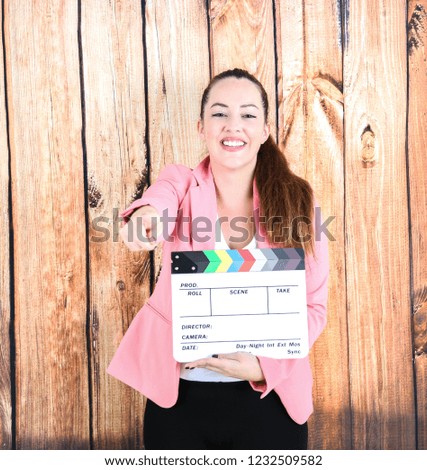 Beautiful young woman holding a clapper board while pointing her finger against a wooden background