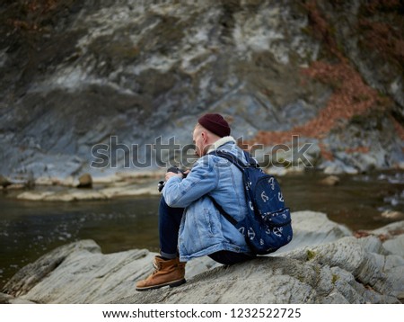 Young man photographer sitting on the rock with his backpack wearing jeans jacket and red hat looking at his camera in the mountains