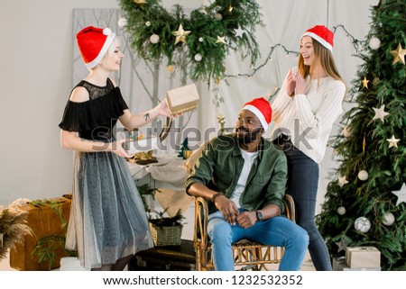 Picture showing group of friends in Santa hats celebrating Christmas at home. Pretty girl in grey skirt gives presents to her friends, African man and Caucasian woman.