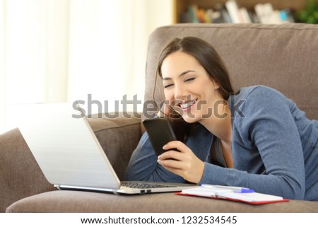 Happy woman using a phone and laptop lying on a couch in the living room at home
