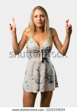 Young blonde woman with fingers crossing and wishing the best. Making a wish. on grey background