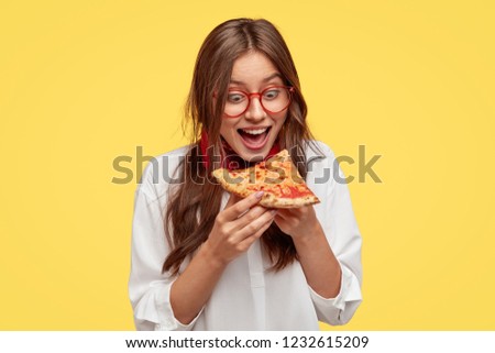 Hungry student opens mouth widely while sees delicious slice of pizza, wants to eat, dressed in white shirt, models against yellow background. Positive woman with junk food. People and eating