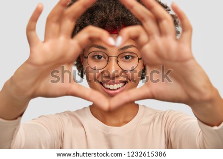 Photo of attractive young woman makes heart shape gesture over face, smiles broadly, demonstrates her love to boyfriend, wears round spectacles, isolated over white background, gestures indoor