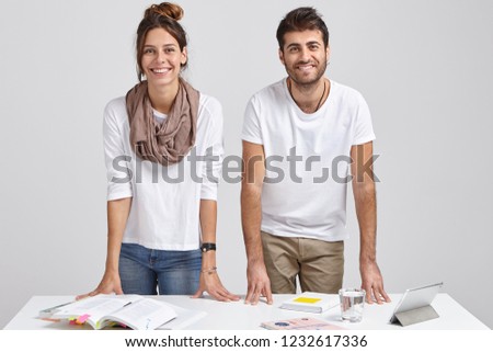 Photo of glad young female and male students lean at table, stand at desktop, learn information, use books and tablet, isolated over white background, solve issues, have brainstroming session