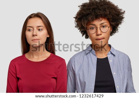 Indoor shot of contemplative mixed race women purse lips, being deep in thoughts, have sad expressions, dressed in casual clothes, isolated over white background. Facial expressions concept. Royalty-Free Stock Photo #1232617429