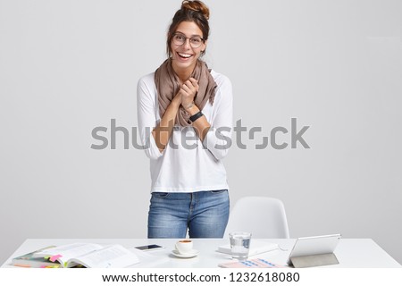 Overjoyed successful lady student makes research alone, uses special application on tablet, stands near white desktop, reads literature, keeps hands together, models against white background