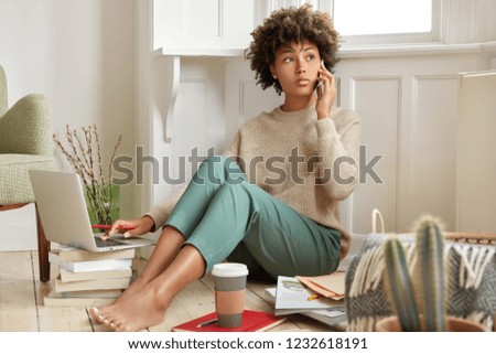 Photo of black woman has telephone conversation, discusses main issues, holds modern cell phone, work with laptop computer, drinks takeaway coffee, reads books, makes course paper, dressed casually