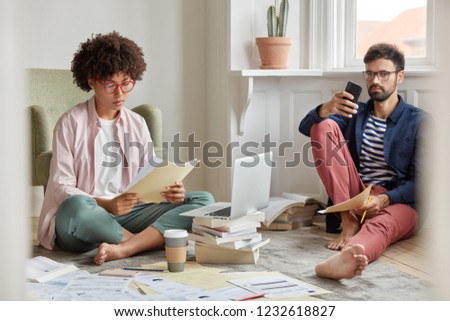 Photo of young stratuppers collaborate together, work at home, sit on floor in living room, think about business investment. Black curly woman in spectacles holds documents, unshaven guy chats online