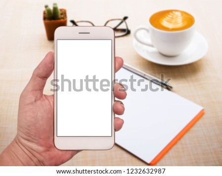 Freelancer's hand holding smartphone with blank white screen on blur office table background.