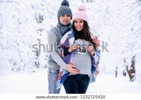 Man holding woman's pregnant belly and the woman holding ultrasound pregnancy picture in her hand while they stand on snowy winter park.
