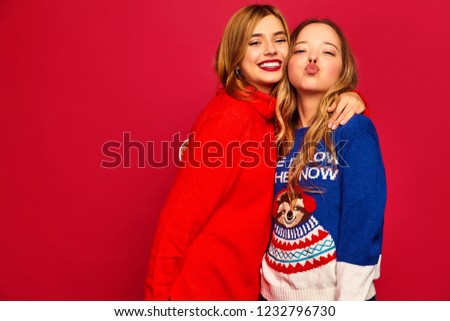 Two beautiful smiling gorgeous girls looking at camera. Women standing in stylish winter warm sweaters on red background. Christmas, x-mas, concept.