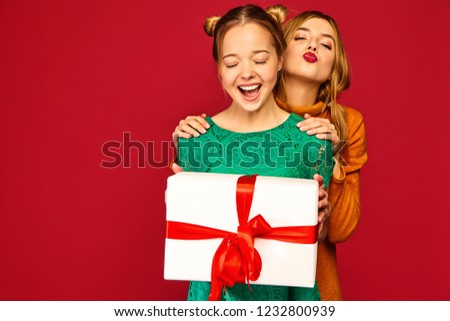 Christmas, x-mas, concept.Two smiling beautiful women in stylish clothes.Girls posing on red background.Model giving her friend big gift box.Having fun,ready for celebration.