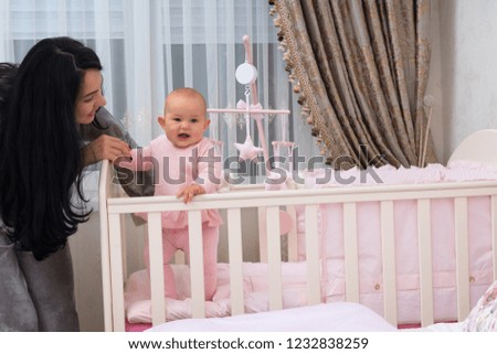 Loving young mother helping her smiling little daughter stand inside her cot in a nursery