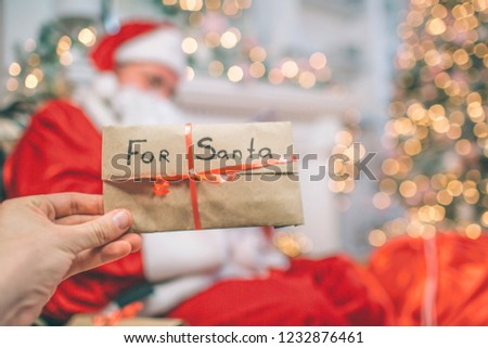 Picture of hand holding letter for Santa. It is covered with red ribbon. There is Santa Claus sitting and holding letters. He reads them.