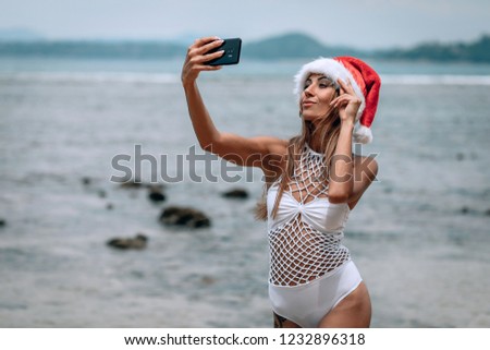 Summer beach vacation girl in santa hat taking fun mobile selfie photo with smartphone. Girl wearing white swimsuit posing for selfie.Trips to warm destinations. Phuket. Thailand.