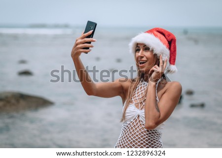 Summer beach vacation girl in santa hat taking fun mobile selfie photo with smartphone. Girl wearing white swimsuit posing for selfie.Trips to warm destinations. Phuket. Thailand.