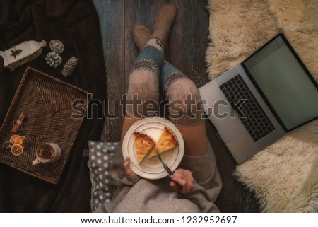 Girl holding plate of christmas cake relaxing at home, drinking cacao, using laptop. Blank screen with copy space for your text message or advertising. Warm cozy home christmas mood. Soft colors.