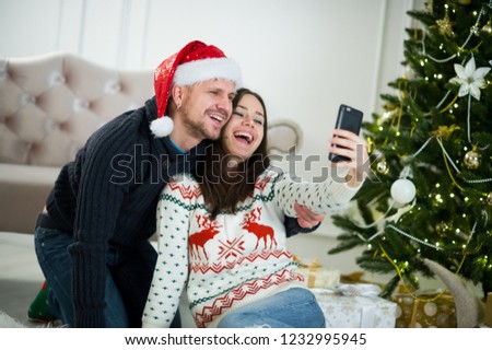 The young couple is photographed against the background of the Christmas tree. The girl takes a picture on the smartphone.