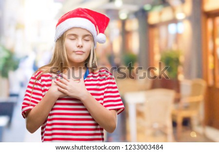 Young beautiful girl wearing christmas hat over isolated background smiling with hands on chest with closed eyes and grateful gesture on face. Health concept.