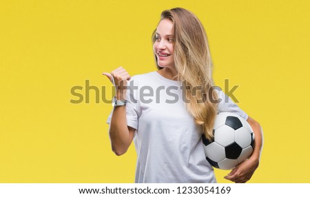 Young beautiful blonde woman holding soccer ball over isolated background pointing and showing with thumb up to the side with happy face smiling