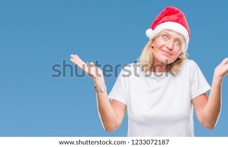 Middle age blonde woman wearing christmas hat over isolated background clueless and confused expression with arms and hands raised. Doubt concept.
