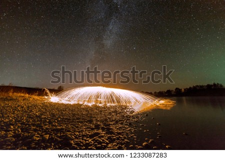 Light painting art concept. Spinning steel wool in abstract circle, firework showers of bright yellow glowing sparkles in fountain form on river bank on blue night starry sky copy space background.