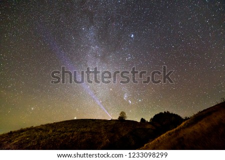 Small silhouette of man with flashlight on dark green grassy hill under beautiful dark blue summer starry sky. Night photography, beauty of nature concept. Wide panorama, copy space background.