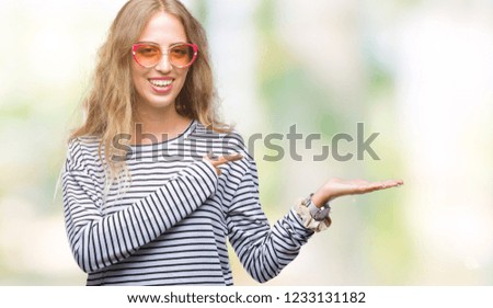 Beautiful young blonde woman wearing sunglasses over isolated background amazed and smiling to the camera while presenting with hand and pointing with finger.