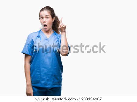 Young brunette doctor girl wearing nurse or surgeon uniform over isolated background pointing finger up with successful idea. Exited and happy. Number one.