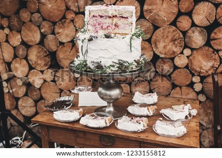 Delicious rustic cake with flowers on silver stand and pieces on plates on log background outdoors. Rustic catering at wedding reception. Barn wedding concept.