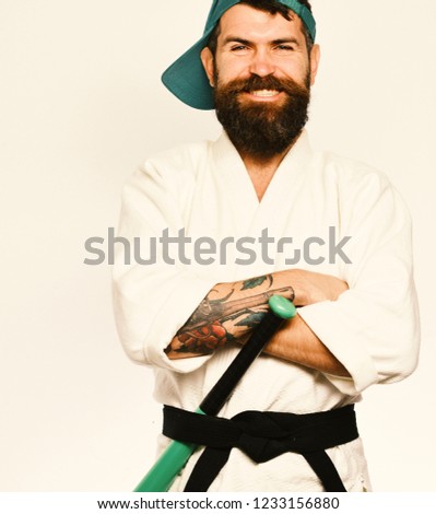 Man with beard in white kimono and green cap on white background. Athlete gets ready to fight. Martial arts concept. Karate man with smiling face puts green baseball bat behind black belt.