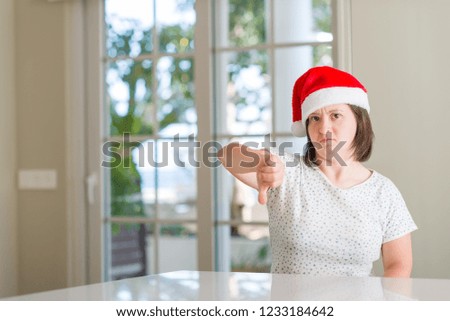 Down syndrome woman at home wearing christmas hat with angry face, negative sign showing dislike with thumbs down, rejection concept