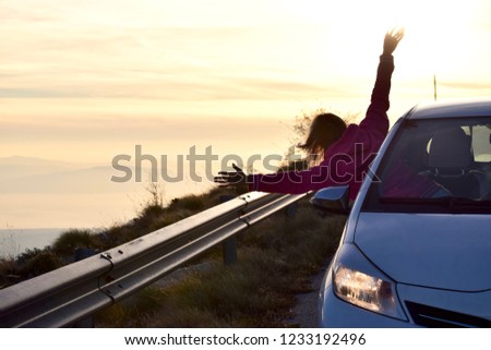Young happy woman sticking out of car window with beautiful sky sunset on background