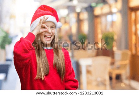 Young beautiful girl wearing christmas hat over isolated background doing ok gesture with hand smiling, eye looking through fingers with happy face.