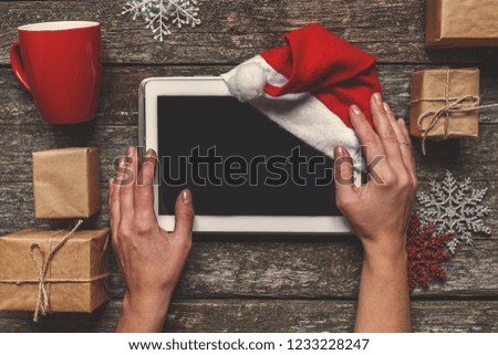 Flat lay top view. Winter Christmas composition. Woman's hands hold a tablet with a Santa hat on a wooden background with festive boxes and New Year's decor.