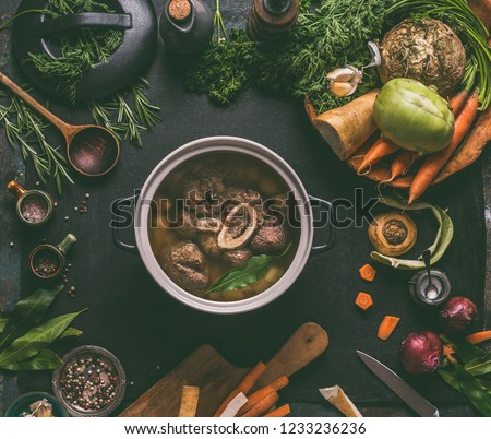 Cooked beef meat shin with bone in cooking pot on dark kitchen table background with low carb vegetables and spices ingredients for soup, top view. Meat broth or stock. Clean low-calorie food concept