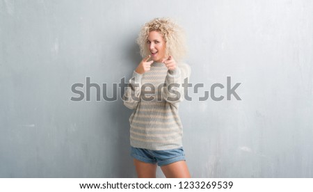 Young blonde woman with curly hair over grunge grey background pointing fingers to camera with happy and funny face. Good energy and vibes.