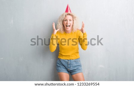 Young blonde woman over grunge grey wall wearing birthday hat celebrating crazy and amazed for success with arms raised and open eyes screaming excited. Winner concept