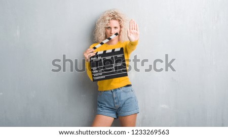 Young blonde woman over grunge grey wall holding calpboard of movie with open hand doing stop sign with serious and confident expression, defense gesture